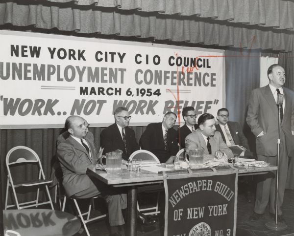 Textile Workers Union (TWU) Director of Research Solomon Barkin stands at a microphone. Next to him, six men are seated at a table with a handmade banner that reads "Newspaper Guild of New York Local No. 3." Behind the table, a banner reads: "New York City CIO Council Unemployment Conference. March 6, 1954. "Work.. Not Work Relief." A caption on reverse of image also identifies some of the seated men: Joseph O'Grady, New York City labor commissioner; John Lotz, Communications Workers of America (CWA); Matthew Guinan, Transport Workers Union of America (TWU); Louis Levine, CIO-CSC [Congress of Industrial Organizations-Civil Service Commission?]. 