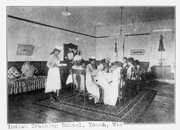 Several women in dresses are seated at a table in a formally decorated room. A woman stands holding a tray at the end of the table. The image appears in a book and includes the captions: "Domestic Science. Dining Room" and "Indian Training School, Tomah, Wis."