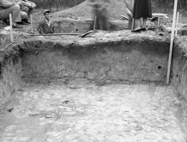 View across an excavation toward men and women standing above and behind it. Men are in another excavated hole behind it. Caption reads: "Excavation in circular mound on R. J. Harker property, Frost Woods, Monona, Dane Co., Wis. A north-south profile through the center of the mound is shown. Excavation Nov. 1, 1947."