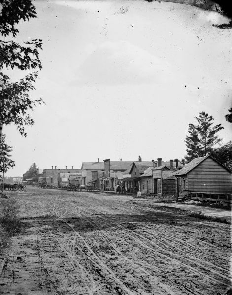 View across and down unpaved street, toward buildings on the right. Many of the storefronts are wood-framed, with two buildings further down the street made of brick. A striped pole on a base is near the boardwalk and porch of a business on the left where men are sitting and standing. Horse-drawn wagons are parked on the left and right side of the street. A sign on one of the buildings reads: "Oakley's Lime House." 
