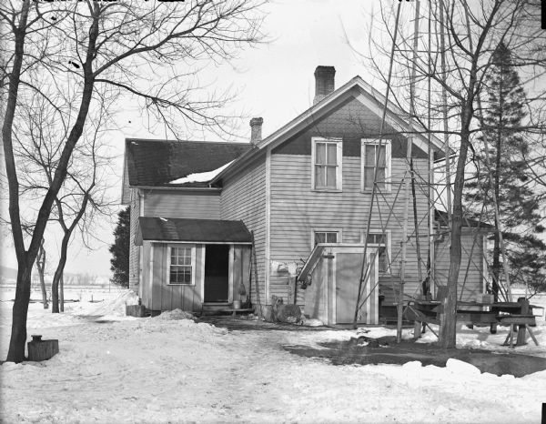 View of a house surrounded by trees. Snow is on the ground, and the base of a windmill is on the right near the house.