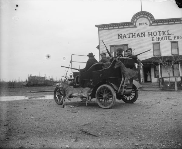 View toward four men riding in a car, with deer fixed to the left running board and the rear. Some of the men are smoking cigars. Behind them is the Nathan Hotel, advertising E[ugene] Houte as proprietor. A woman is standing in the hotel doorway. In the background are railroad cars on a railroad track loaded with logs.