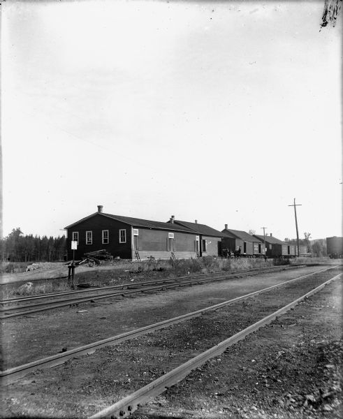 View across two sets of railroad tracks toward several buildings. Two men are in a horse-drawn carriage in front of the buildings near railroad cars on a siding. The buildings have narrow doors with shutters, and short ladders against the building below. Caption reads: "Scandinavia, Wis. c1900-1905. Scandinavia Produce Co. with tracks of GB&W RR [Green Bay & Western railroad] in foreground."
