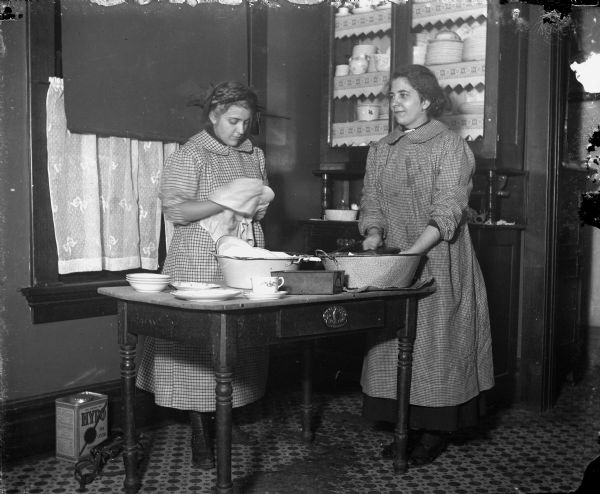 Two women are standing at a table in a kitchen, with one woman drying a dish, and the other washing dishes in a large bowl. Dishes and pans are stacked on the table, and a glass fronted cabinet behind them holds dishes.