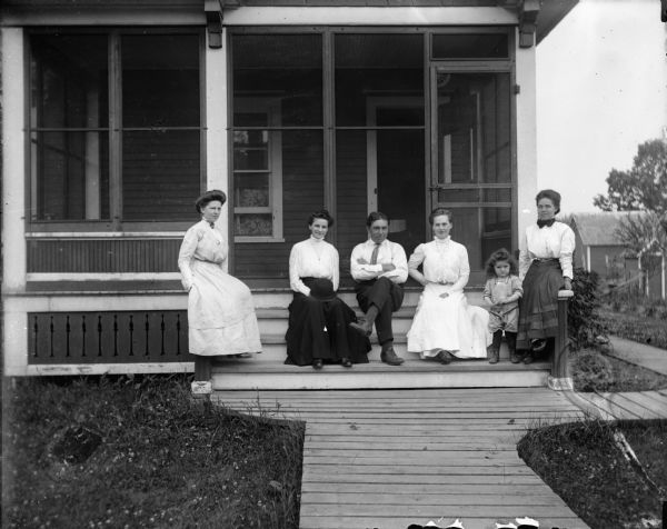 Four women, one man and a child are posing on the front steps of a screened-in porch. A wooden walkway leads to the porch, and over and around the right side of the house. Outbuildings are in the background. Caption reads: "Gluths house with party."