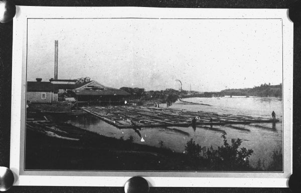 Slightly elevated view of a large building complex along a river, with a raft of logs in the water in front of it. A man is standing at the end of the raft on the far right. Caption reads: "Daniel Shaw Lumber Co., Wis. Mill with raft in foreground."
