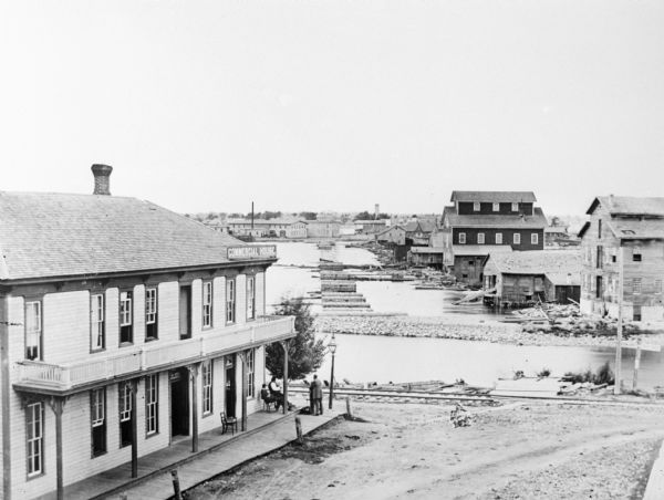 Elevated view of several buildings, with bridge and railroad tracks, and the Fox River. In the foreground on the right is the Commercial House, with three men sitting and standing on the porch and board sidewalk at the corner of the building near the river. Industrial buildings and boathouses are along the water, and on the opposite shoreline are industrial buildings. Caption reads: "The Manufacturing Plants along the Old Bridge in DePere in 70's. They consist of machine shop, saw mill, grist mill and wooden pump works."