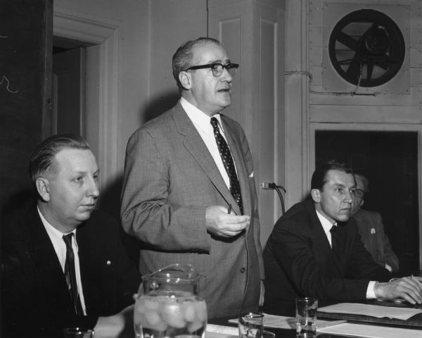 A man stands and gestures with a pen in hand. Other men are seated on either side of him. Caption reads: "General President William Pollock addressing meeting of the Greater Toronto Textile Joint Board. L to R: Jack Phillips, newly elected joint Board president; Pollock; Paul Swaity, Canadian director; Ray Ruggles, Toronto [customs?] agent."