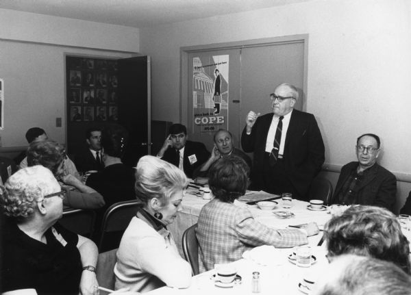 A man is standing and gesturing at a table. Men and women are seated at tables nearby. A poster on the door reads: "for good government and just laws give a buck to COPE, Committee On Political Education, AFL-CIO." Caption reads: "Keynoting 1968 institute, then Gen. Pres. William Pollock stresses need for political activity to insure that the gains at the bargaining table are not 'lost in legislative halls.'"