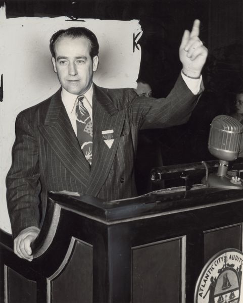 A man stands at a podium, gesturing. A seal on the front of the podium reads: "Atlantic City Audito[rium]." Caption identifies him as William Pollock, general secretary-treasurer of the Textile Workers Union of America [TWUA].