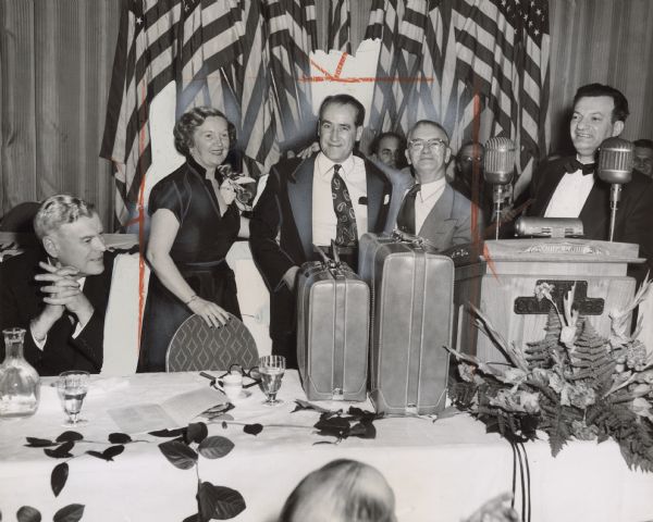 Two men and a woman are posing while standing behind a table. One of the men has his arm around the other. A set of luggage is on the table in front of them. A man on the right is standing behind a podium that reads: "The Commodore." A fourth man is sitting on the left. Caption reads: "SET OF LUGGAGE was Philadelphia Local 80's gift to [TWUA general secretary-treasurer William] Pollock. Duffin Magee (right) made presentation. Mrs. Pollock rounds out the trio."