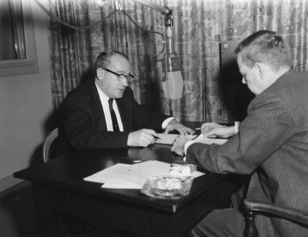 Two men are seated at a small table facing each other with a microphone suspended between them. Caption reads: "TEXTILE PROBLEMS make the airwaves. Left, General President William Pollock is interviewed by IUD [sic] Pipeline's James Toughill...."