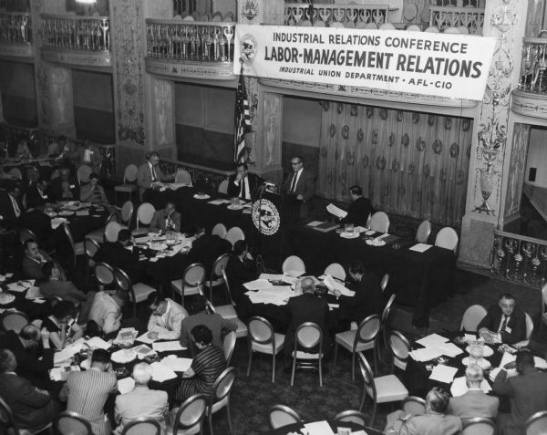 Elevated view of a man speaking at a podium and groups of people sitting at tables around the room. A banner suspended from a balcony reads: "Industrial Relations Conference. Labor-Management Relations. Industrial Union Department - AFL-CIO." Caption reads: "DIXIE STORY of brutality and lawlessness against workers related by TWUA's William Pollock at IUD's first industrial relations conference in Washington proved real eye opener to the delegates from already well-organized areas."