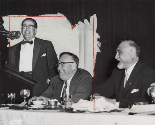 A man is standing at a podium and smiling. Two other men are seated near him and also smiling. Caption reads: "LIGHT MOMENT at testimonial dinner finds the guest of honor taking a ribbing from Executive Vice President William Pollock (left)--and getting a bang out of it."