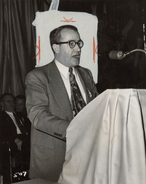 A man stands at a podium and speaks into a microphone. Two men are seated in the background. Caption reads: "ORGANIZING'S EVEN MORE IMPORTANT, Executive Vice President William Pollock reminded conference delegates. He called for a stepped-up drive in central Jersey."