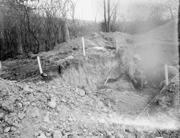 Men are working in a hole dug in the ground, which is roped off with stakes. Caption reads: "Excavation in circular mound on R. J. Harker property, Frost Woods, Monona, Dane Co., Wis. Nov. 1, 1947."