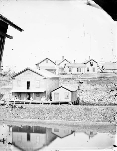Slightly elevated view across a river toward a large building with Greenwood Mills written on the side. The mill is reflected in the river in the foreground. A man at the mill is leaning in the open doorway under the roof of the loading dock, and sacks are piled near him on the left and right. An open door above the man has belt driven machinery inside. On a hill behind the mill is a long fence and other buildings, including a church with a dome on top of a tower in the background on the right. 