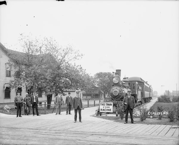 Several men pose on a station platform in front of a locomotive and railroad car at the end of the railroad tracks. A man standing in the center is wearing a hat that reads: "Brake Man." A sign in front of the train reads: "This Train For Racine." Taken in Corliss, in the Mount Pleasant township of Racine County.