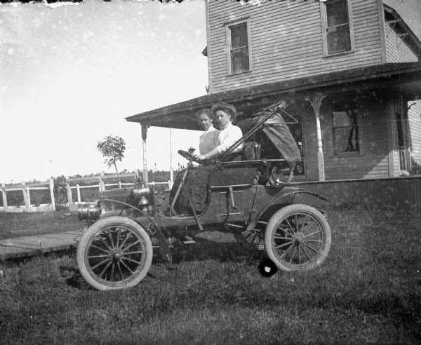 Two women are seated in a car in front of a house. A small fence and a board sidewalk is between the car and the house in the background on the left.