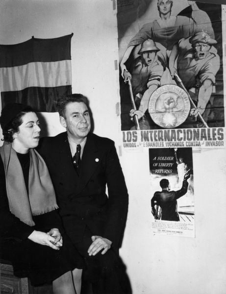 A man and woman are seated in front of a flag and next to two posters. Caption reads: "War Bride and Groom Back From Spain. New York, N.Y. — Captain Hans Amlie, of the Washington-Lincoln Battalion of the Abraham Lincoln Brigade in Spain, and his bride, the former Milly Bennett of San Francisco, newspaper woman, who were married recently at Albacete, Spain, on a "certificate of death", when it was not possible to produce original birth certificates, are shown. They were forced to make use of the device which permits a dying person to be wed, although neither was ill at the time. Mrs. Amlie, who has been in China, Russia, Spain and other European countries for the past seven years, met her husband while serving as head of the Valencia Press Bureau for the Loyalists. He was in Army service and had been wounded twice in the war. The couple are pictured at the Friends of the Abraham Lincoln Brigade Headquarters here."