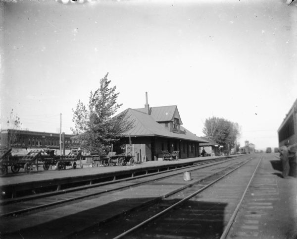Angled view across two sets of railroad tracks toward a depot, which has numerous carts lined up on the platform. On the far right a man is standing next to a railroad car on railroad tracks. Caption reads: "Billings, Montana (?), c.1915-1930. Railroad station of the NP [Northern Pacific], CB&Q [Chicago, Burlington and Quicy]."
