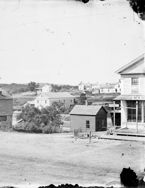 Slightly elevated view toward several buildings. Fences run between them, marking yards. Caption reads: "River Falls, Wis. View showing Greenwood Mills and other buildings. Looking from Main Street & Maple." A small sign on the front of the large building on the right reads: "Hudson 10 Mi. Prescott 1.12 Mi."