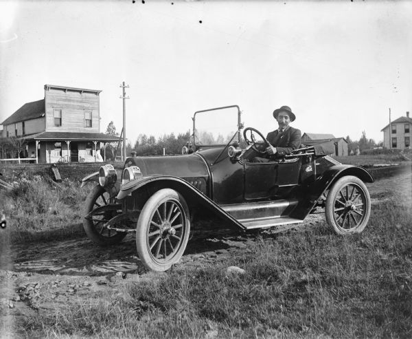 A man poses behind the wheel of a car in the foreground. In the background on the left two women are on the porch of a building with a false front. In the background on the right is a large building and two small outbuildings.