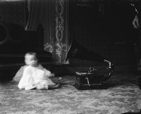 A baby (blurred from motion) is sitting on the floor next to a phonograph, in front of a bed. An adult's hand is visible on the far right. Caption reads: "Madison (?), Wis. c.1910-1915. Baby and phonograph."