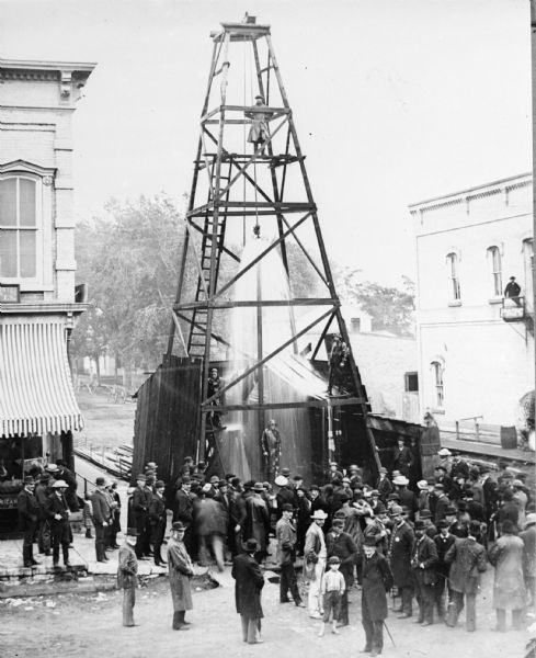Elevated view of a crowd of people standing near an artesian well. Men wearing long coats and hats to protect them from the water are standing on  and under the tower structure. Commercial buildings are on either side of the well. Caption reads: "First Artesian Well in DePere, Still in Use. Drilled in 1885 - natural pressure furnished water for city for many years through mains."
