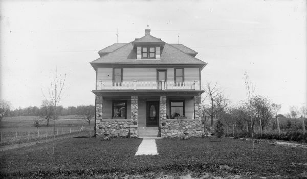 View across lawn toward the front of the house, with a stone front porch and a balcony above. A fence is along the right and left bordering the yard. Fields and a hill are in the background.