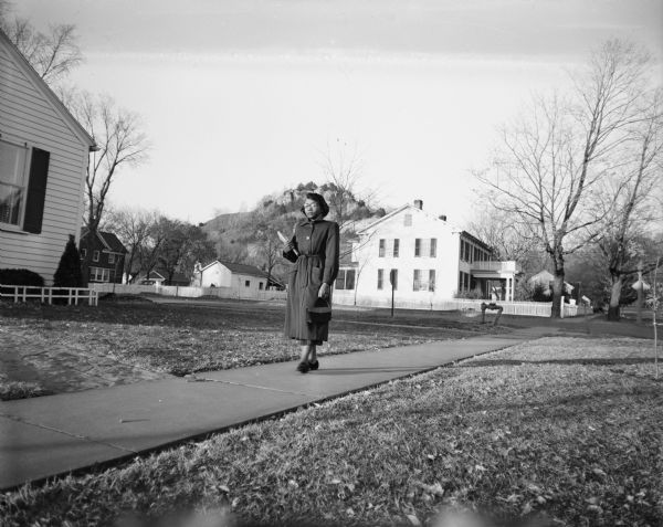A woman, Mrs. Rebecca Wyatt, is walking along a sidewalk. She is the first black school teacher in Cassville. Mrs. Wyatt moved to Wisconsin from Paris, Tennessee; she was hired as a commercial teacher for the local high school. As reported in the paper at the time, the school district did not know prior to her arrival that she was not white. She was welcomed warmly, however, especially after the community learned that her husband was stationed with an Army engineer unit in Korea.