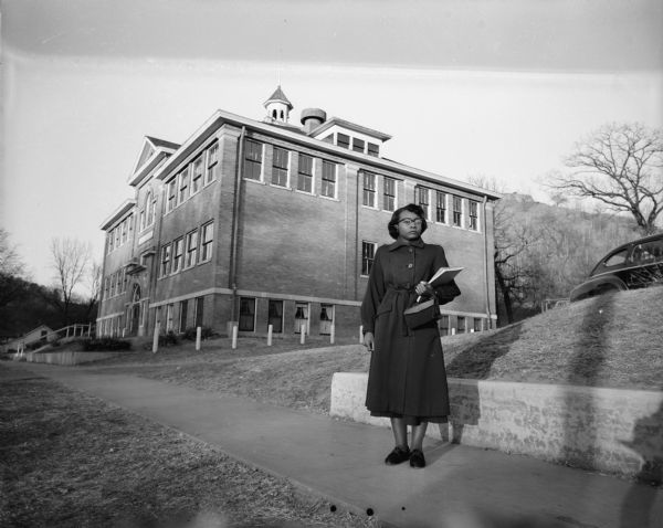 A woman is standing on a sidewalk outside a school, holding a notebook. This is Mrs. Rebecca Wyatt, the first black school teacher in Cassville. Mrs. Wyatt moved to Wisconsin from Paris, Tennessee; she was hired as a commercial teacher for the local high school. As reported in the paper at the time, the school district did not know prior to her arrival that she was not white. She was welcomed warmly, however, especially after the community learned that her husband was stationed with an Army engineer unit in Korea.