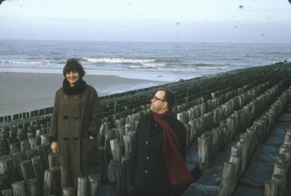 German film critics Enno Patalas and Frieda Grafe are seen at the beach in Knokke-le-Zoute, Belgium.  The two are on a groyne (a wooden and cement barrier) and are standing apart from each other.  Patalas looks up at Grafe.

