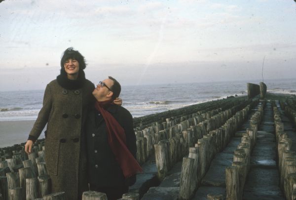 German film critics Enno Patalas and Frieda Grafe stand on a groyne (a wooden and cement barrier) on the beach in Knokke-le-Zoute, Belgium.  Grafe is higher then Patalas and has her arm around his neck as he looks up at her.  