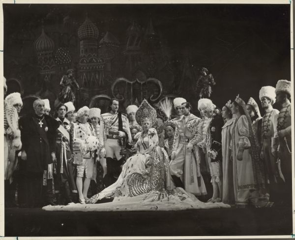Mae West, as Catherine the Great, sits surrounded by military officers and diplomats in a scene from the play "Catherine Was Great".  West wears a lavish fur-trimmed gown and a large headpiece.