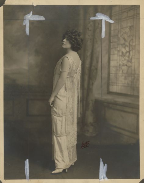 Publicity photograph of Mae West circa 1912.  West wears a sleeveless dress with beaded overlay.  She stands with her back to the camera and looks over her shoulder.  Her hair is dark with big curls.  There are crop marks on the photograph.