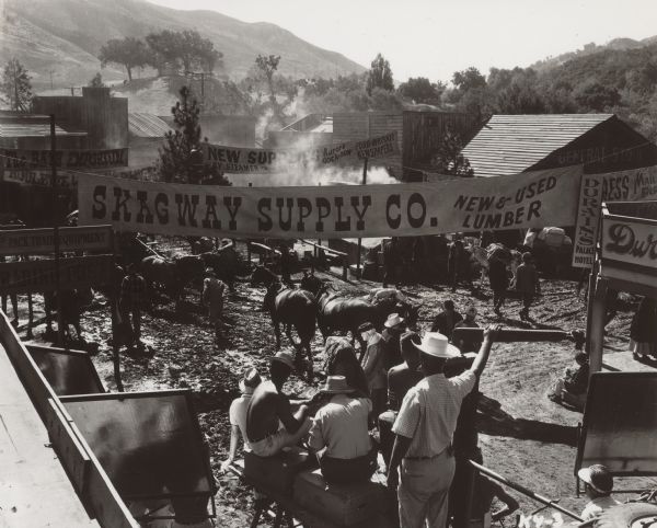 Photograph of a street scene being filmed for the 1960-1961 Ziv-TV series "Klondike". Large banners hang across the street.  A camera and crew are in the foreground filming people and horses in front of them.
