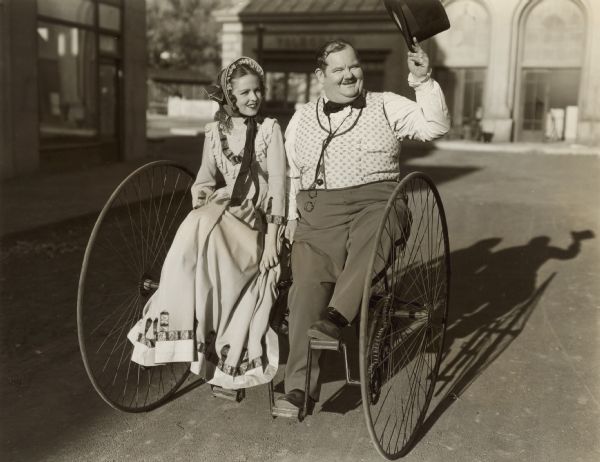 Oliver Hardy and Alice Koerner sit on a two-person bicycle for a publicity photograph for the 1939 film "Zenobia."  Hardy holds a top hat in one hand.  The pair and the bicycle are on a street.