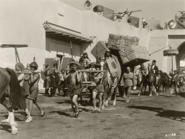 Cast members of the 1938 film "The Adventures of Marco Polo" carry material using shoulder yokes and rides horses loaded with straw boxes.  Lighting equipment can be seen on top of the walls in the background.