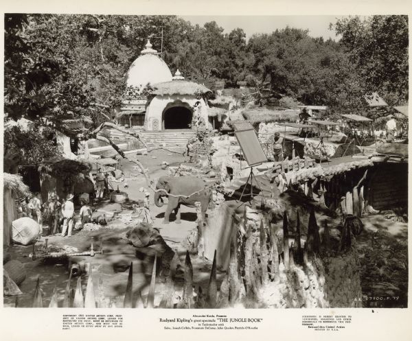 Photograph of the 1942 film "The Jungle Book."  A domed building with an arched entry way with thatched roof is seen in the background.  An elephant, crew members, and lighting equipment is seen in the foreground.