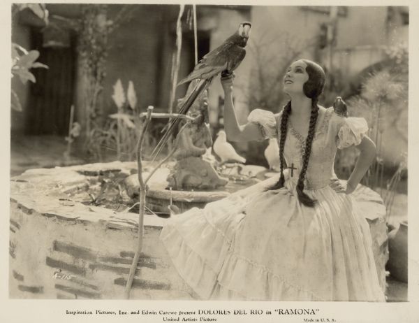 Dolores del Rio appears as the title character in the 1928 film "Ramona".  She wears a dress, a crucifix around her neck, and long braids while sitting next to a fountain with a parrot perched on one hand.