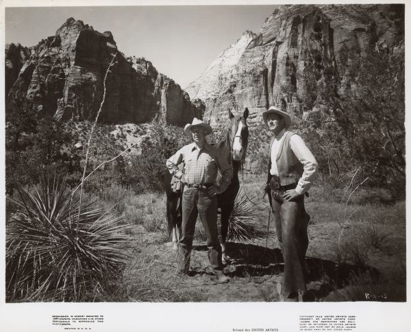 Actor Joel McCrea and another man stand with a horse in front of two large mountains.  Both men are dressed as cowboys and stand with their hands on their hips.  The photograph is a publicity photograph for the 1947 film "Ramrod".