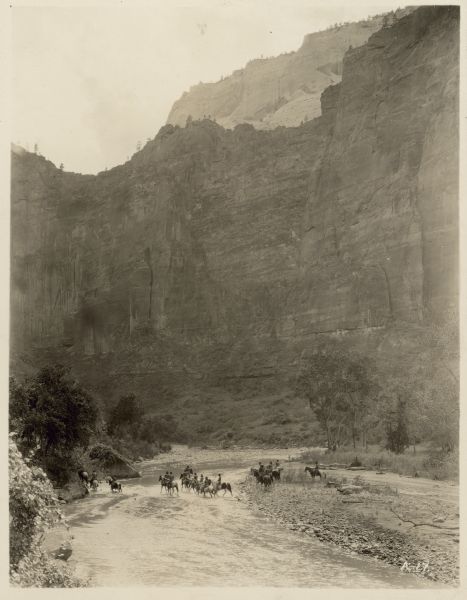 Men on horses, dressed as Native Americans, cross a river in a scene from the 1928 film "Ramona".  The river is at the base of a mountain cliff.