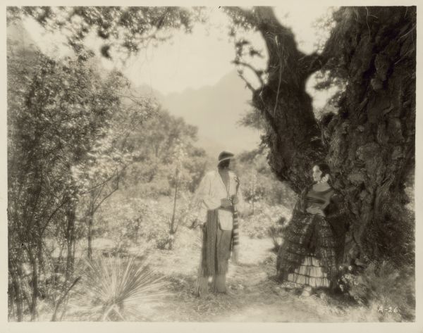 Dolores del Rio and Warner Baxter stand beneath a tree in a scene from the 1928 film "Ramona".  Baxter portrays Alessandro, a Native American, and del Rio is Ramona, a part Native American and part Mexican.