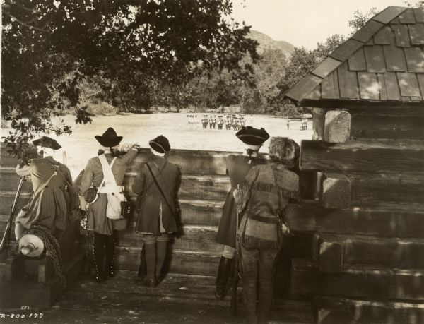 Photograph taken on the set of the 1936 film "The Last of the Mohicans" of British soldiers peering over a wooden wall.  They are looking at a group of French soldiers outside Fort McHenry.