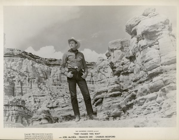 Actor Joel McCrea poses in front of an impressive cliff while shooting the 1948 film "Four Faces West."  He is dressed as a cowboy, has his thumbs hooked onto his jean pockets and has a pistol tucked into his waistband.