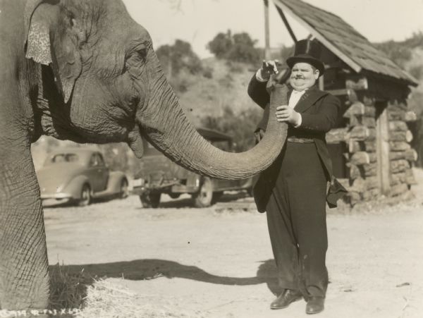 Oliver Hardy, as Doc Tibbett, feeds Queenie the elephant, who plays the title character in the 1939 film "Zenobia."  Hardy wears a tuxedo and top hat and holds the elephants trunk while holding a bit of food above it.