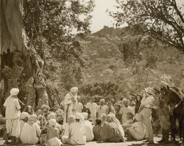 A group of people sit and stand while listening to a man speak in a scene from the 1942 film "The Jungle Book."  All the men wear turbans while the women wear scarves on their heads.  A woman standing on the far right, next to a horse, wearing pants and a hat takes a photograph of the group.