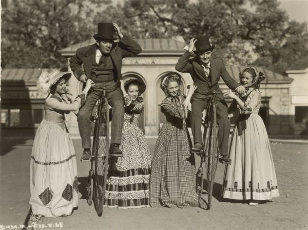 Ruth Fitzgibbon, Marjorie Kane, Marie Barde, and Alice Koerner hold onto Pat Lane and Jack Egan as they sit atop penny-farthing bicycles on a street.  The group all appear in the 1939 film "Zenobia."