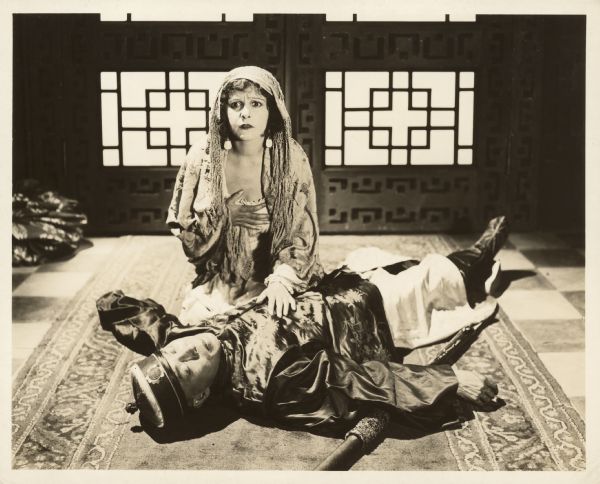 Norma Talmadge, as San San, kneeling next to the body of man, possibly her father, in Chinese clothes in a scene from the 1918 film "The Forbidden City".  She holds her hand to her chest and looks frightened.
